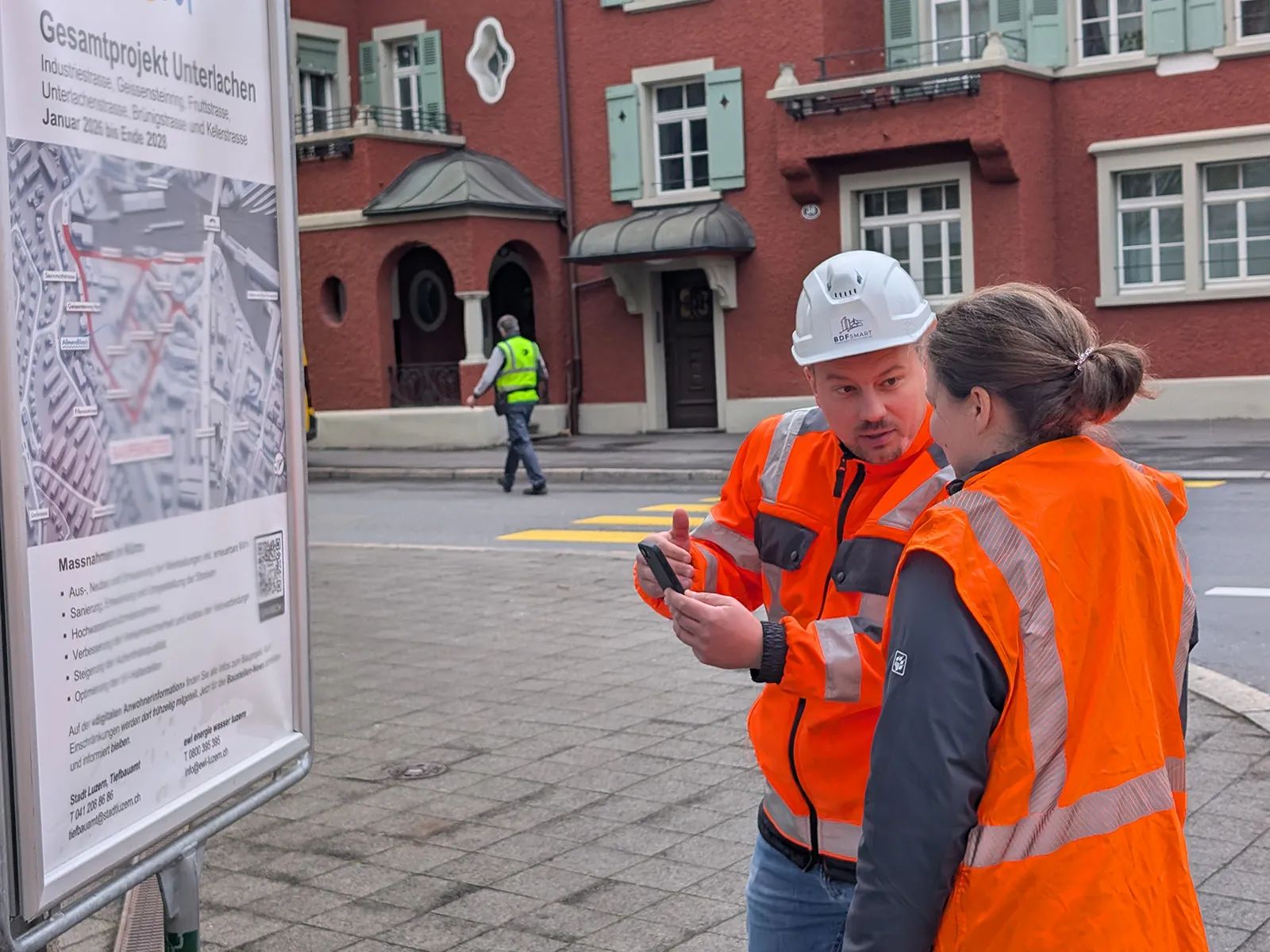 Two project members in orange high-visibility clothing discuss the construction information on a smartphone in front of the «Gesamtprojekt Unterlachen» information board in Lucerne.