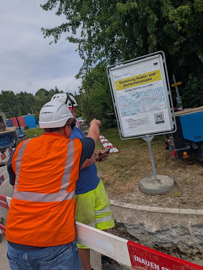 Two project members in helmets, one in an orange high-visibility vest, in front of the Maur information board for the renovation of Auwisstrasse and Dorfacherstrasse; one points at the board, the other holds a smartphone.