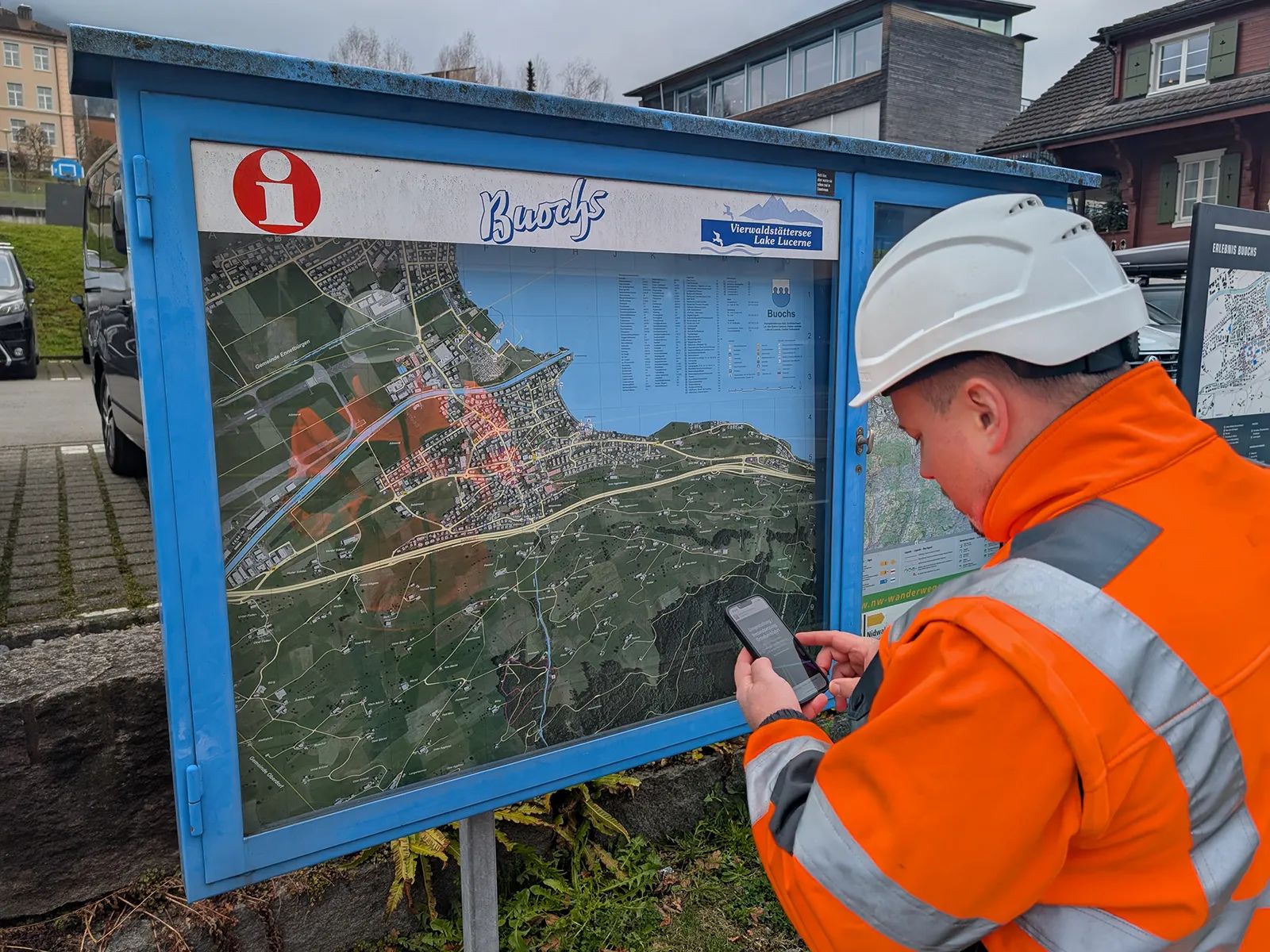 A project member in a helmet and orange high-visibility jacket opens the construction information on a smartphone next to the Buochs village map board.
