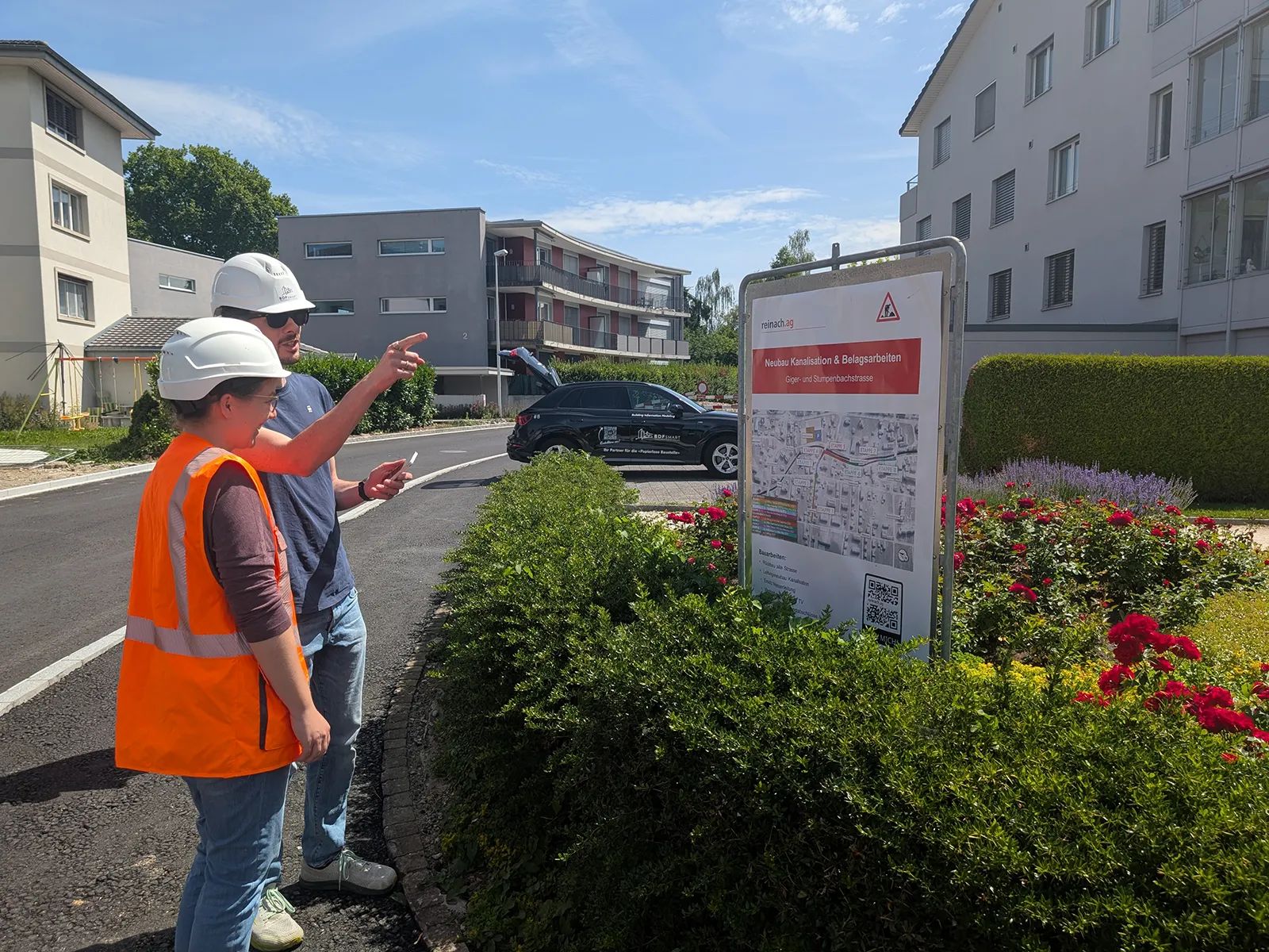 Two project members in helmets in front of the information board for the sewer and paving works on Gigerstrasse and Stumpenbachstrasse in Reinach; one points along the street.