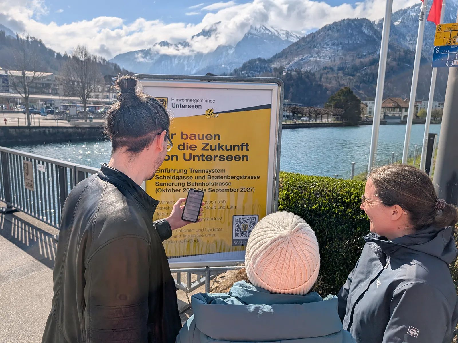 Three people on the waterfront in Unterseen reading the municipal information board on the separate sewer system Scheidgasse–Beatenbergstrasse; a man opens the construction information on a smartphone, lake and mountains behind.