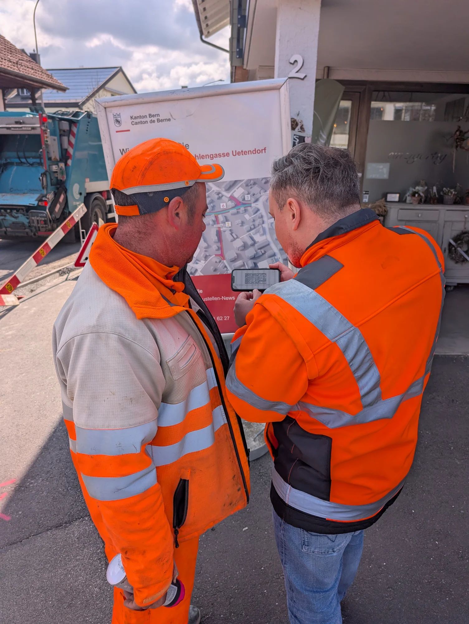 Two construction workers in hi-vis clothing open the construction site information on a smartphone in front of the site board on Hohlengasse in Uetendorf.