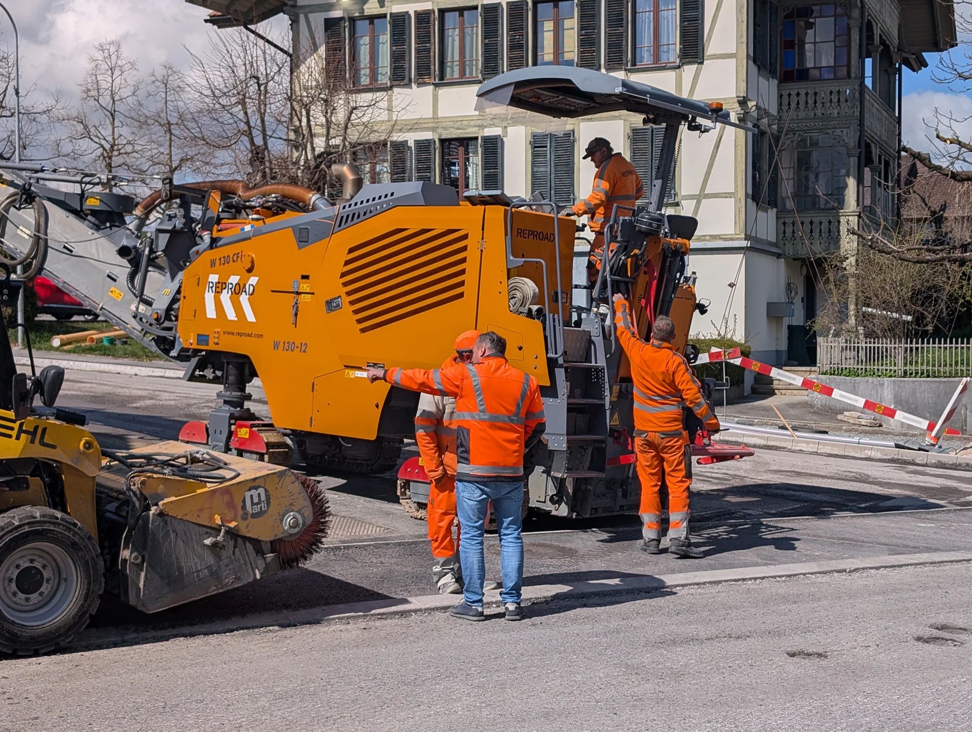 A road milling machine and a sweeper remove the old surface in front of a historic house; workers in hi-vis clothing guide the machine.