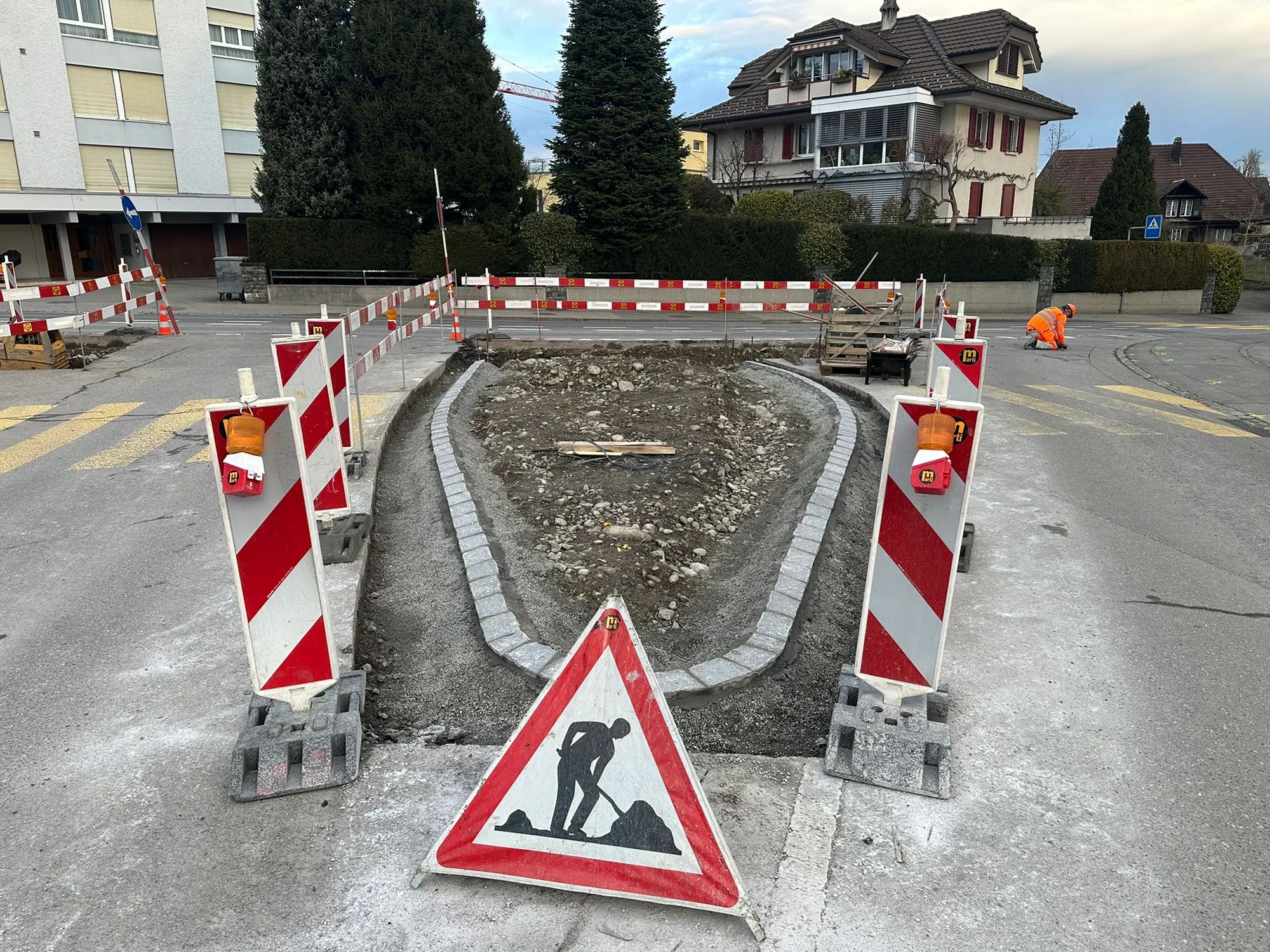 Traffic island under construction with new kerbs, barrier beacons and a roadworks sign.