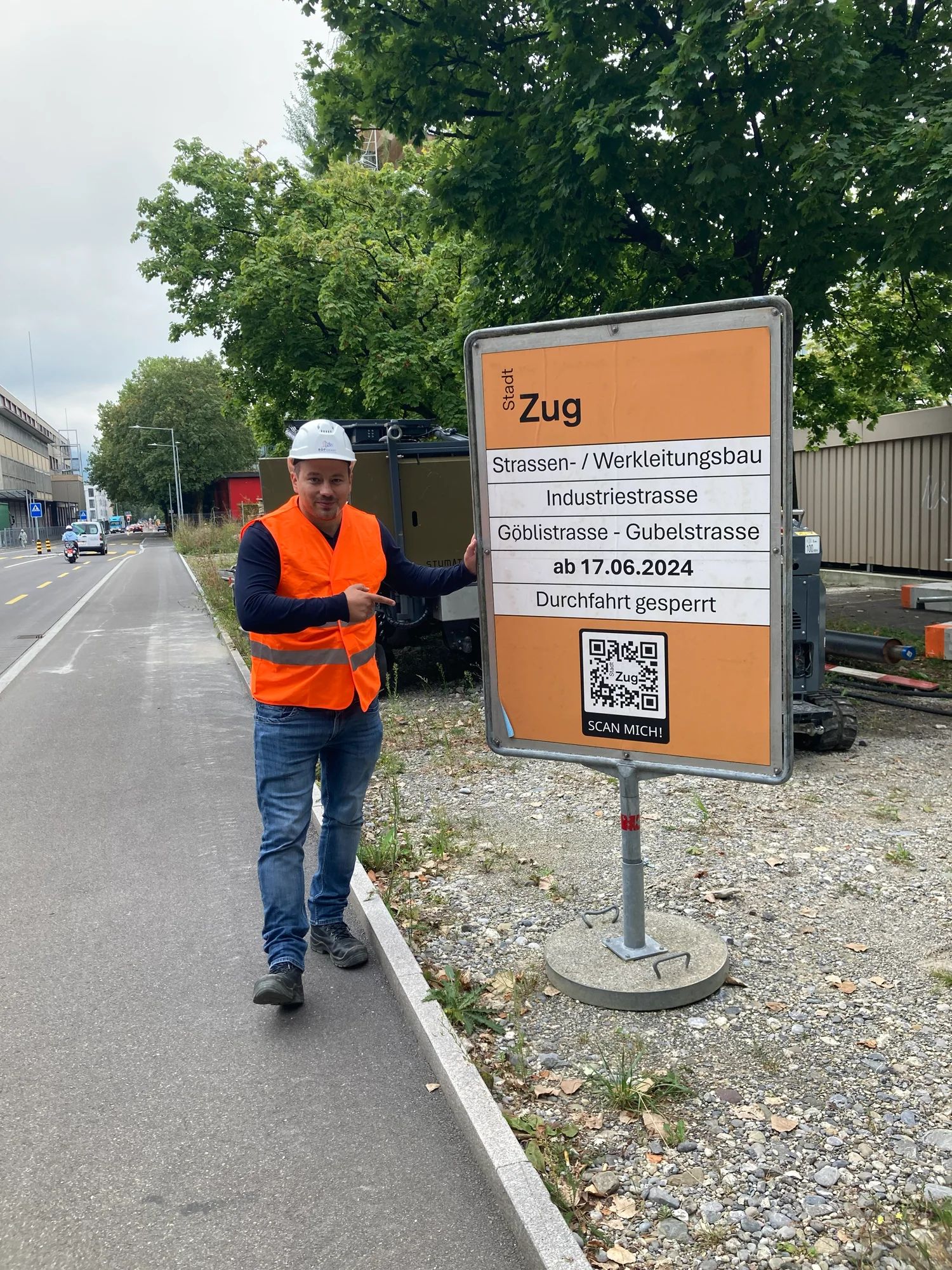 A project team member in a hi-vis vest points to the City of Zug site board for the Industriestrasse road and utility works, with QR code.