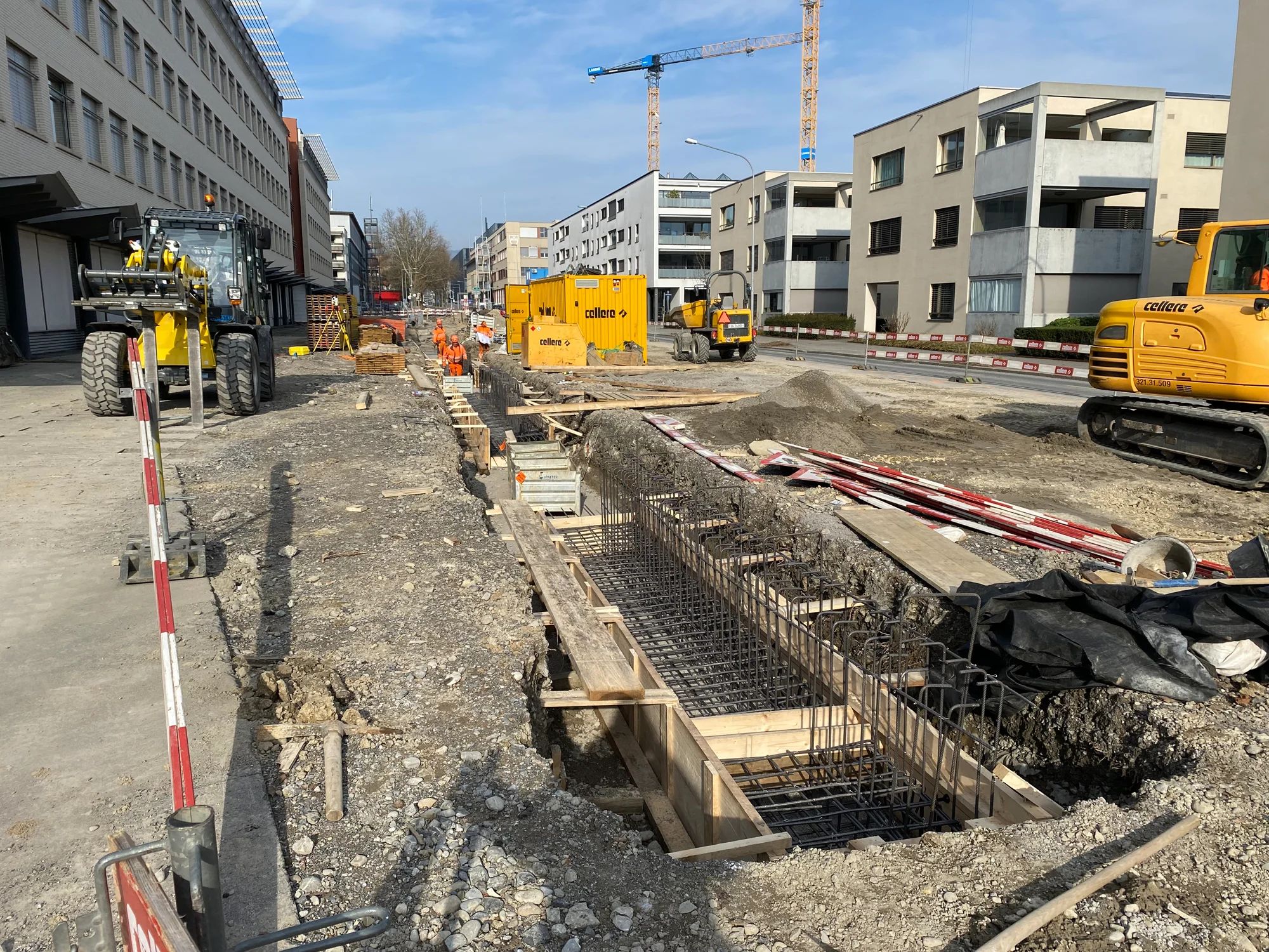 Open excavation with reinforcement next to the carriageway; construction machinery, cranes and apartment buildings behind.