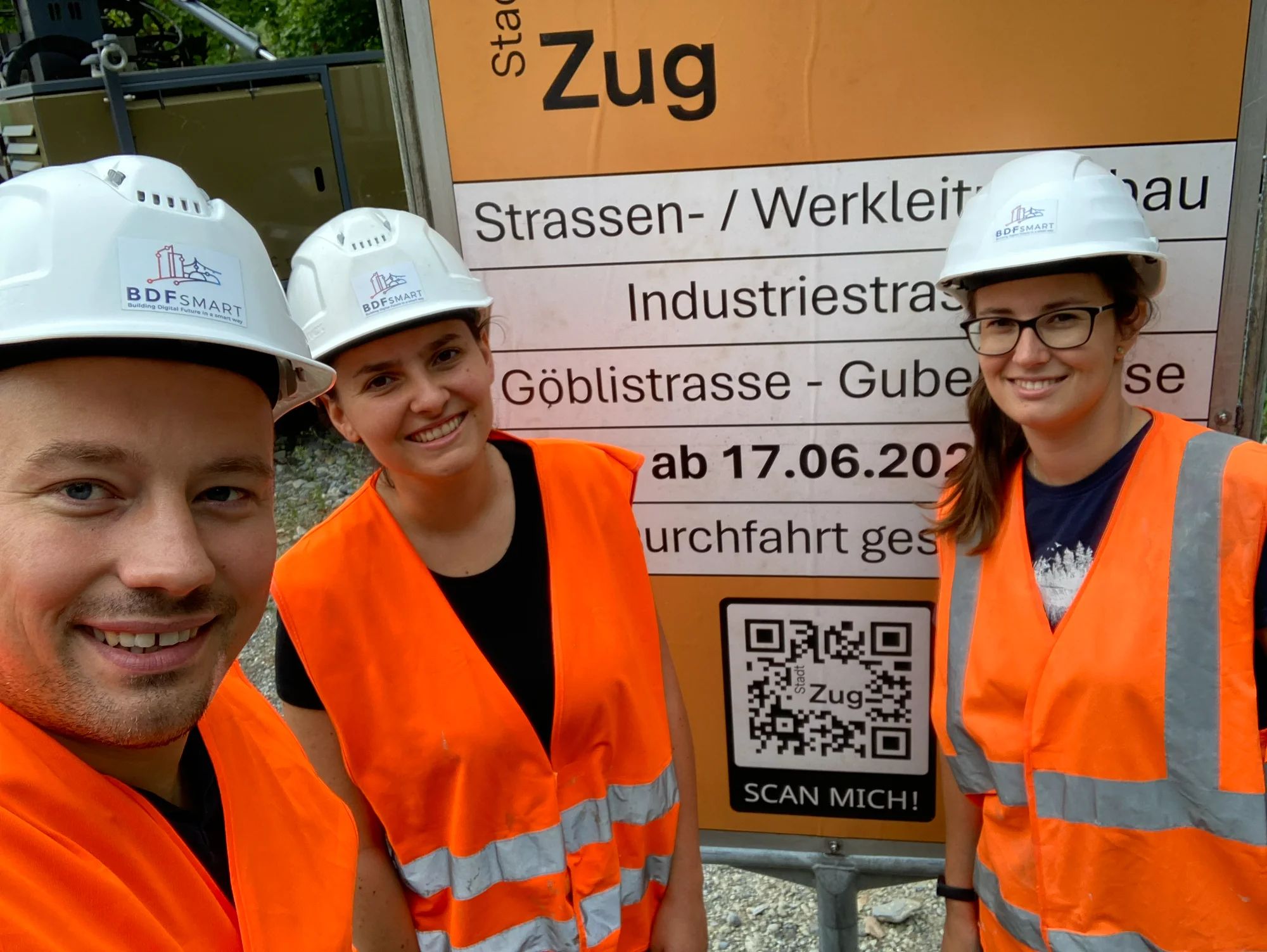 Three project team members in BDFsmart helmets and hi-vis vests in front of the site board with QR code.