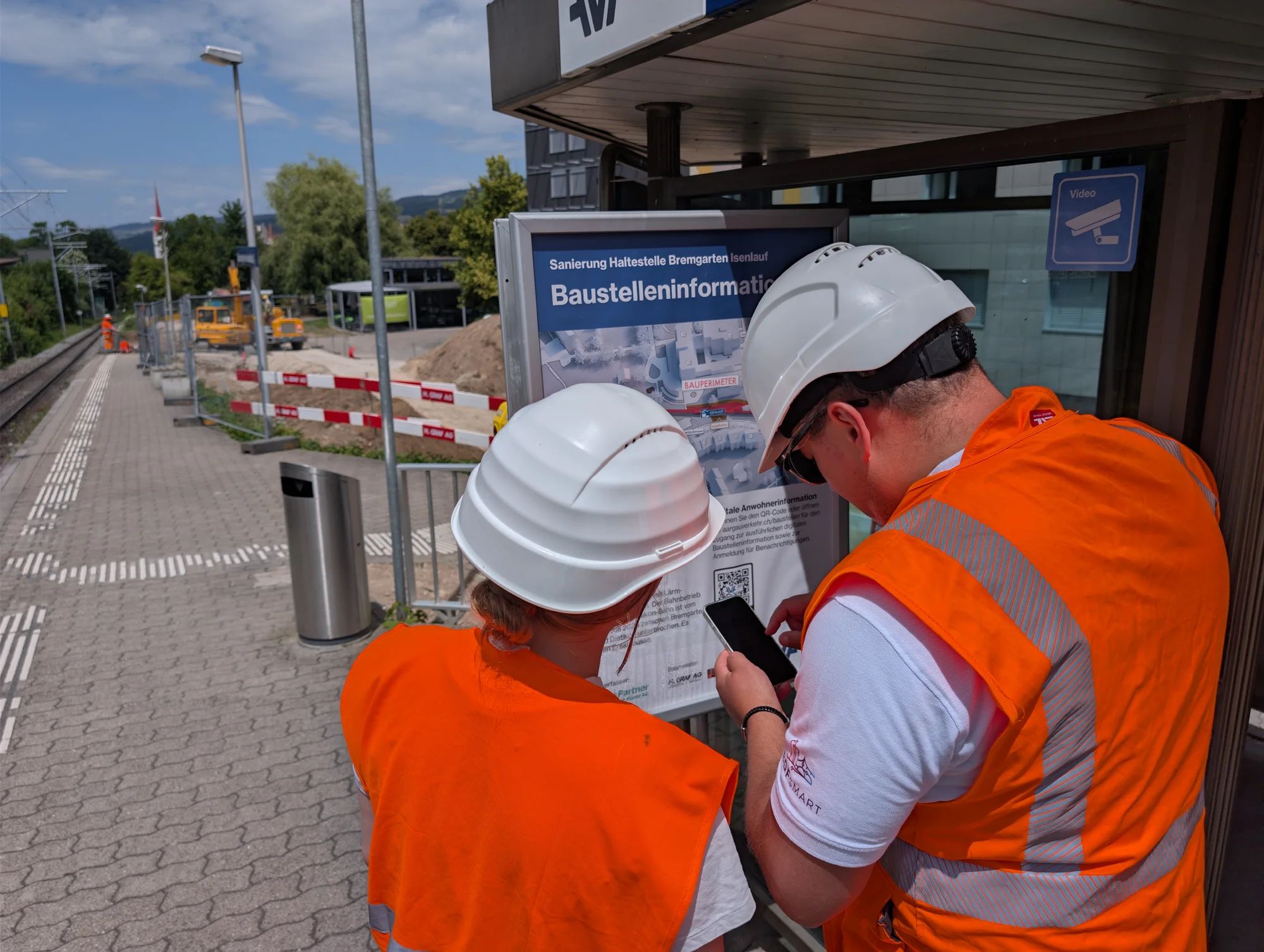 Two project team members in helmets and hi-vis vests scan the QR code on the construction site information board at the Isenlauf stop in Bremgarten with a smartphone.