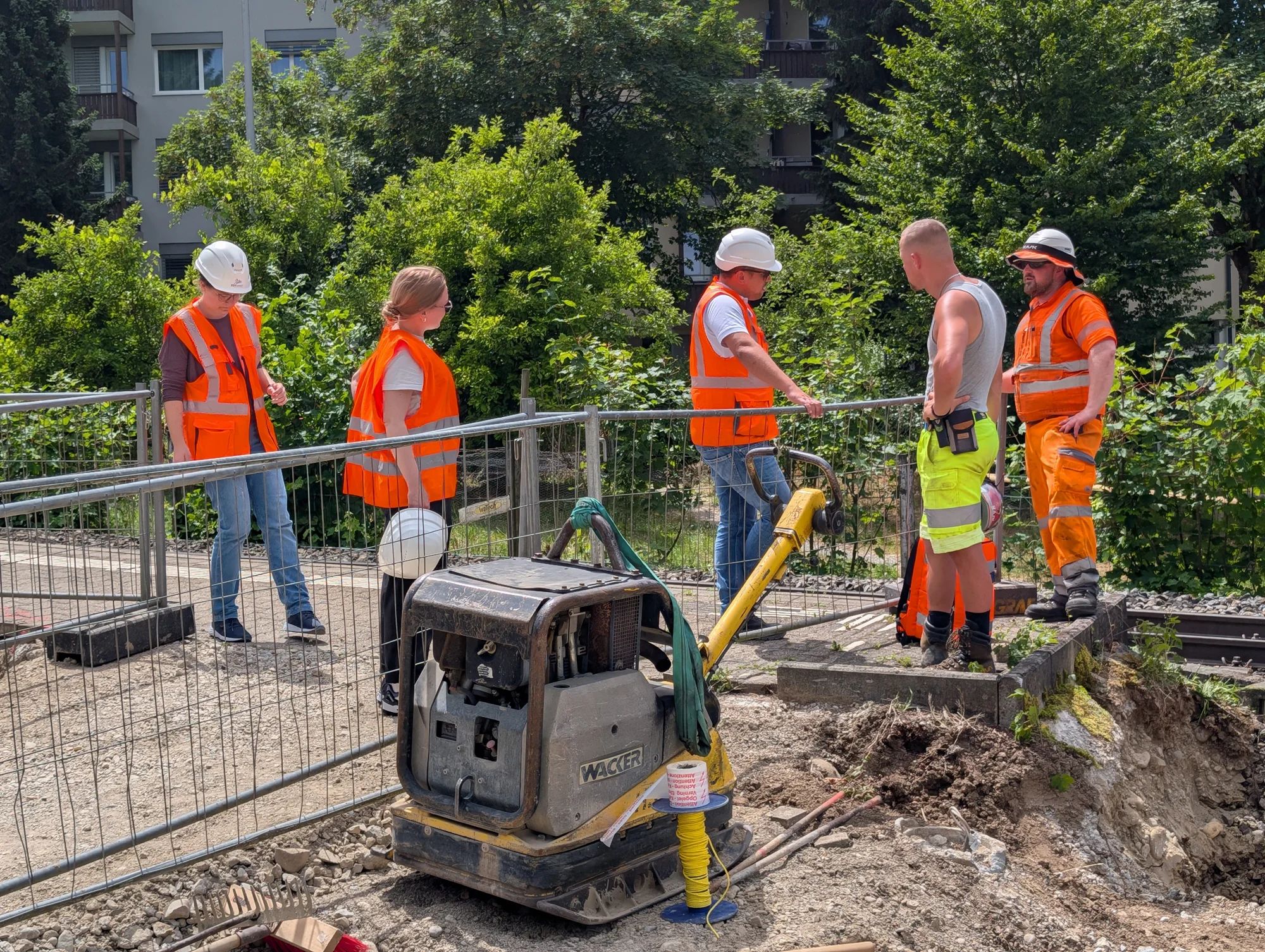 Project team members and construction workers talk at the site fence next to the track; a plate compactor in the foreground.