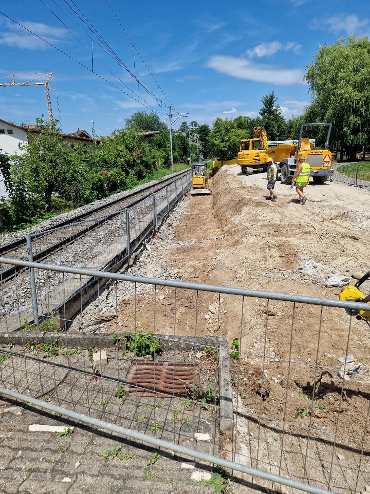 Construction track along the railway line with machinery and workers; the site fence in the foreground.