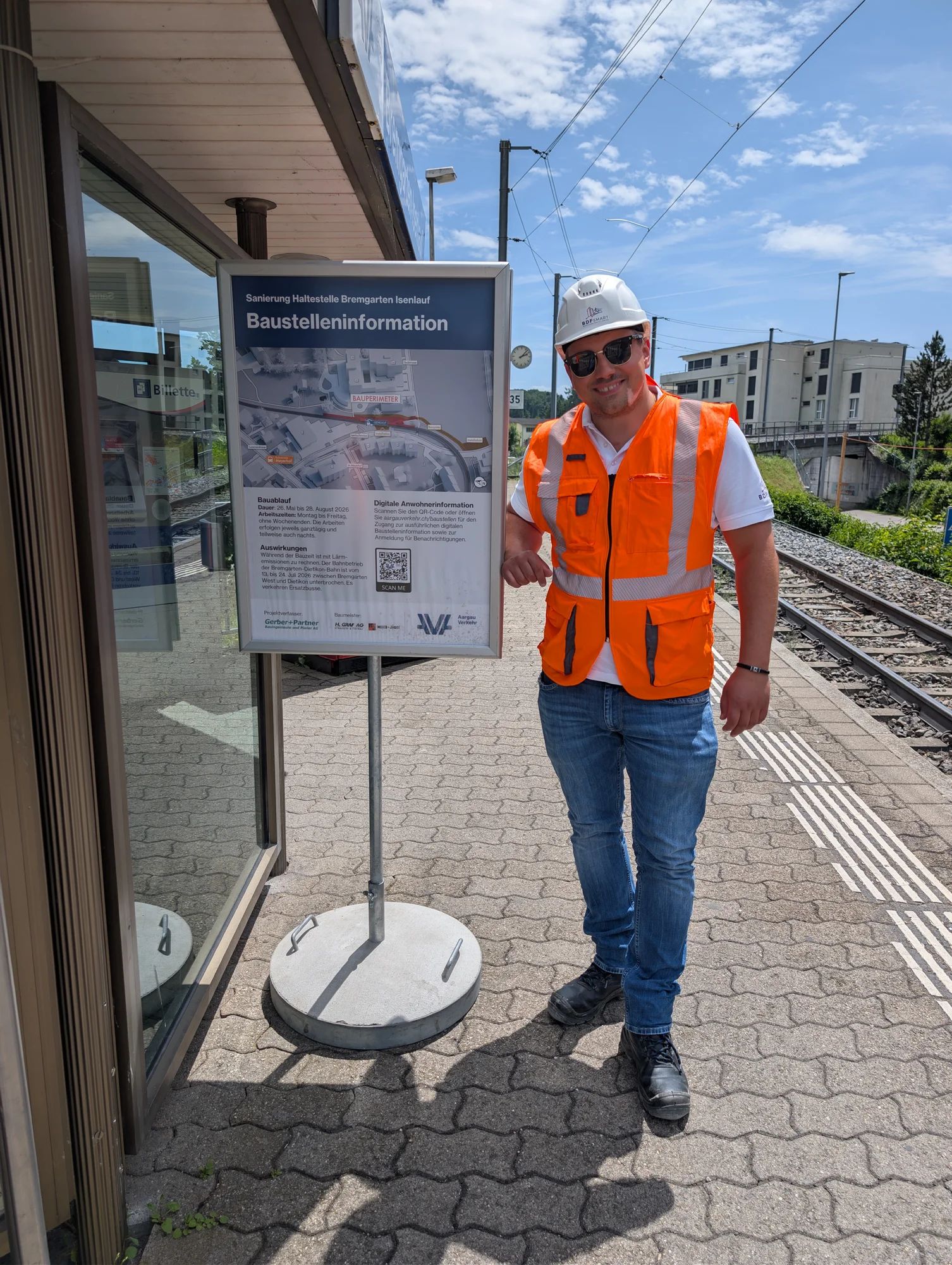 A project team member in a helmet and hi-vis vest on the platform next to the construction site information board.