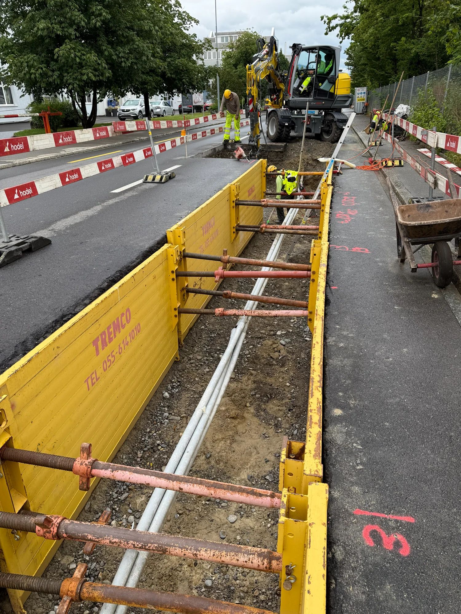 Shored utility trench with conduits along Knonauerstrasse; behind it an excavator and workers in hi-vis clothing.