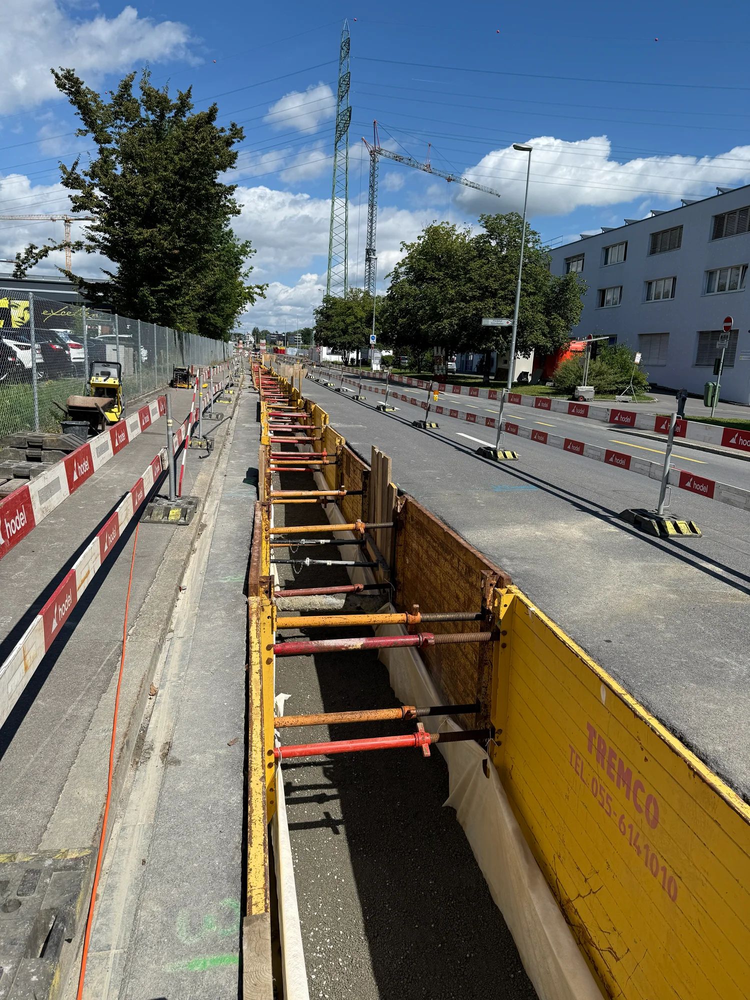 Long trench secured with yellow shoring panels next to the cordoned-off carriageway; a pylon and cranes in the background.