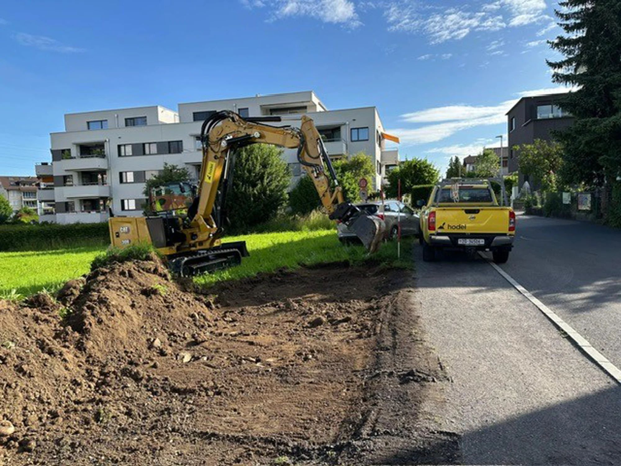 A mini excavator digs on a verge beside the road, with a pick-up truck next to it.