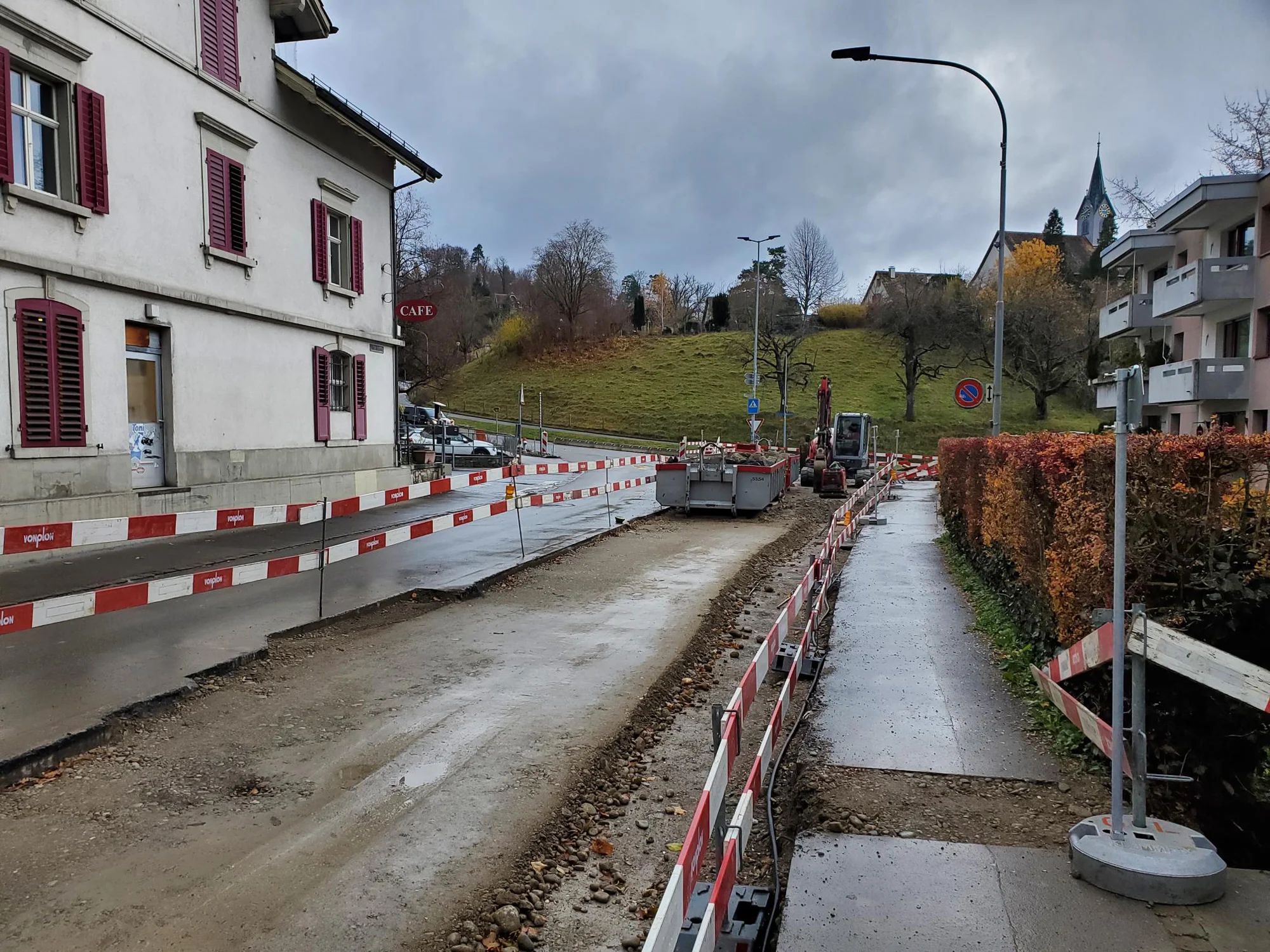 Cordoned-off roadside on Sihlwaldstrasse with an excavated trench beside the footpath; an excavator and the church tower in the background.