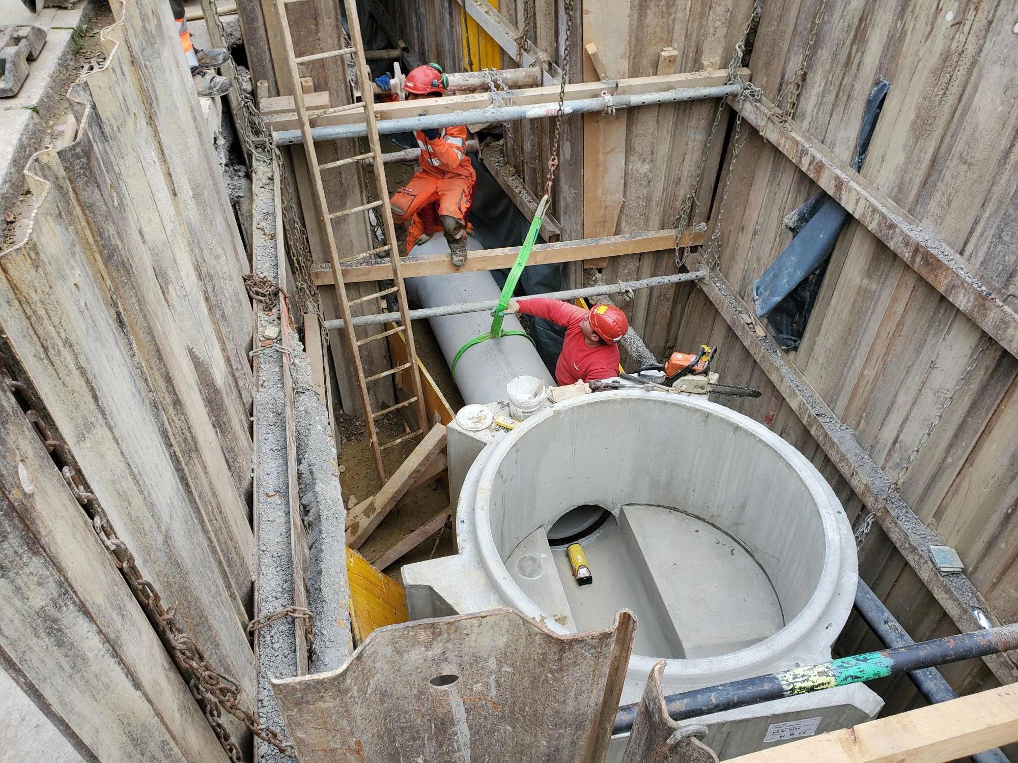 Workers install a concrete manhole in a shored excavation pit.