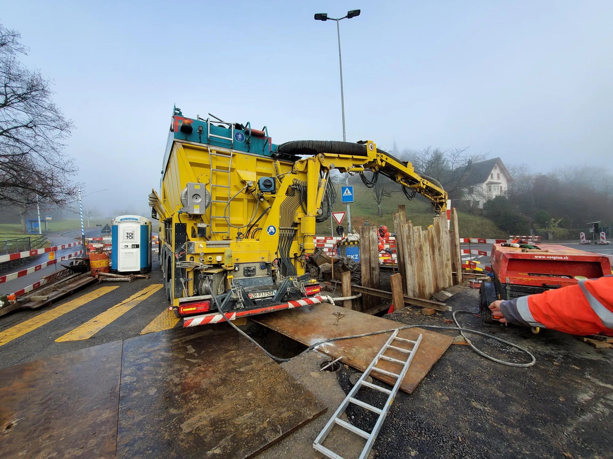 A yellow vacuum excavator at an open pit on the cordoned-off junction.
