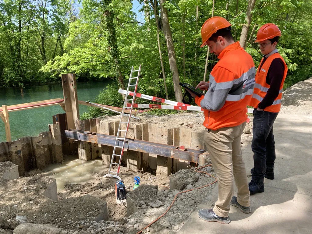 Two specialists in high-visibility clothing check the project on a tablet at an excavation on a riverbank