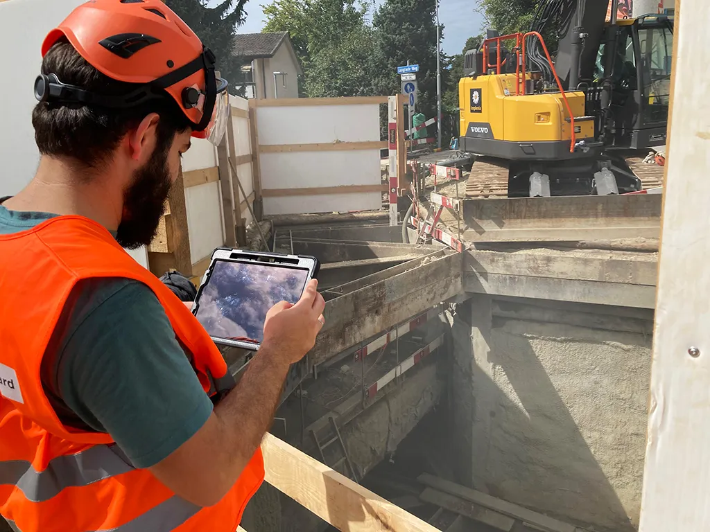 Specialist in a helmet and orange high-visibility vest checking the model on a tablet at the shored Wehrenbachhalde excavation; excavator behind.