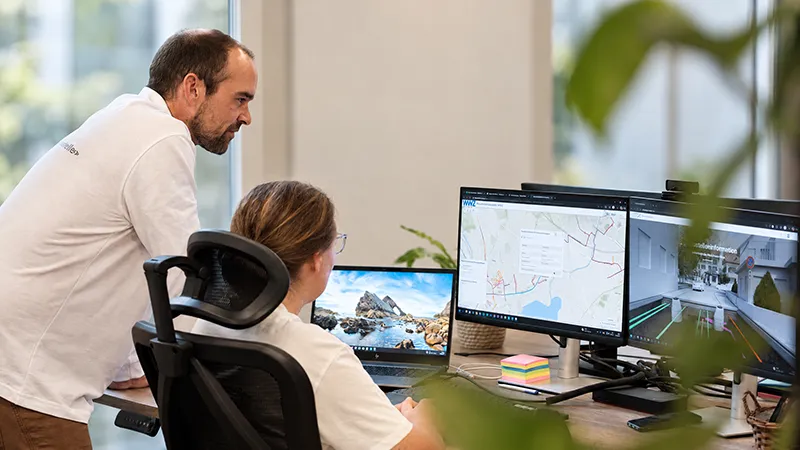 An employee looks over a colleague's shoulder at her office workstation; the screens show a map of a utility network and a 3D street view