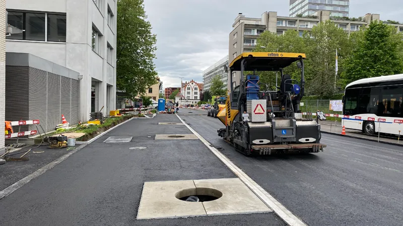 Industriestrasse during surfacing: a paver on the carriageway, a bus alongside and a new manhole in the foreground.