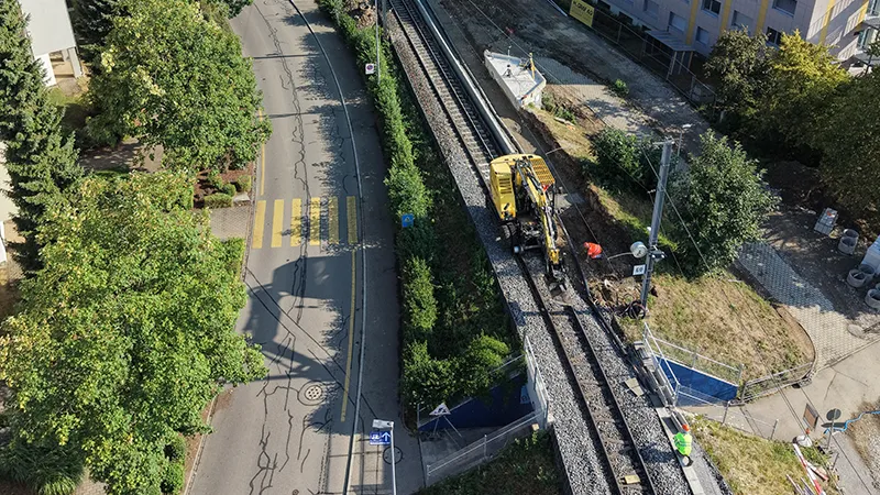 Aerial view of the site: a yellow construction machine on the track next to a residential street with a pedestrian crossing.